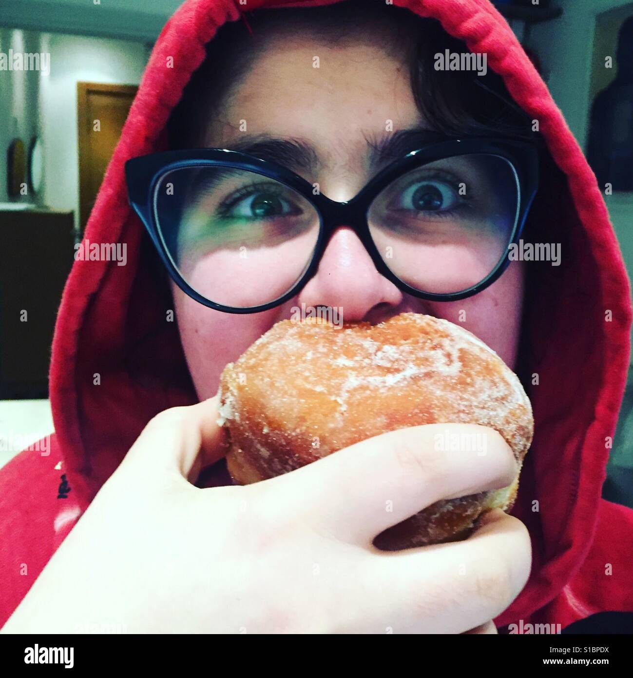 Mädchen mit großen Gläsern rot Hoodie und rosa Wangen beißen in einen großen Stau-Donut Stockfoto