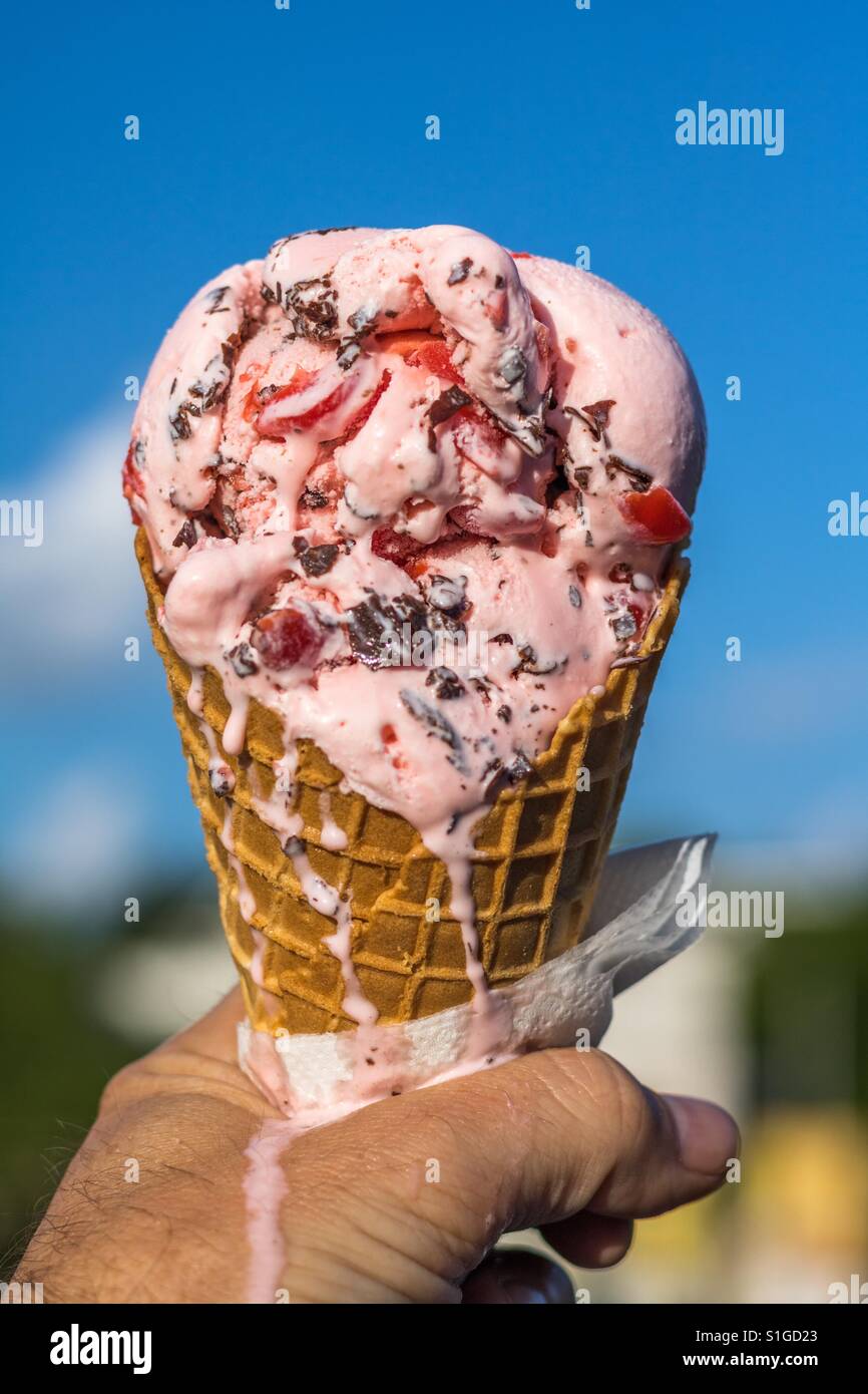 Vertikale Foto des Schmelzens Cherry chocolate Chip Eis in einer Waffelkegel gegen ein strahlend blauer Himmel mit Wolken und einige Bäume im Hintergrund unscharf gehalten Stockfoto