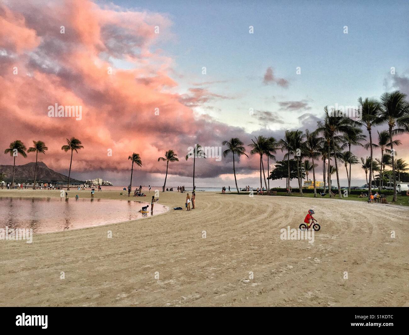 Waikiki Beach und Diamond Head mit rosa Gewitterwolken Overhead bieten schöne Kulisse kleiner Junge Reiten Fahrrad auf Sand von tropischen Palmen gesäumten Lagune, Oahu, Hawaii Stockfoto