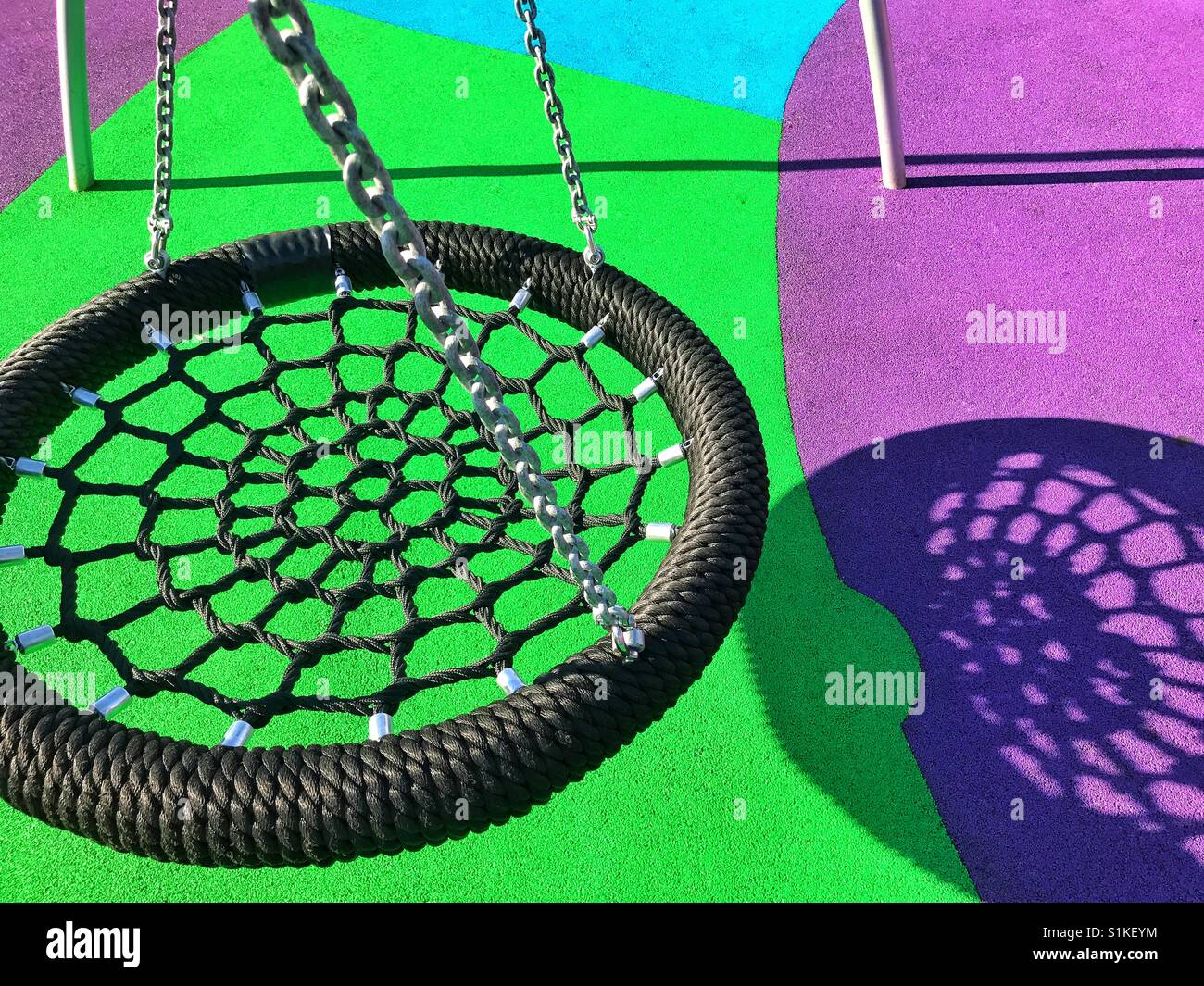 Eine neue Schaukel wirft einen interessanteren Schatten auf den bunten Boden in einen Kinderspielplatz. Eine Grafik mit interessanten Farben & Formen. Bildnachweis - © COLIN HOSKINS. - Smartphone-aufgenommenes Stockfoto