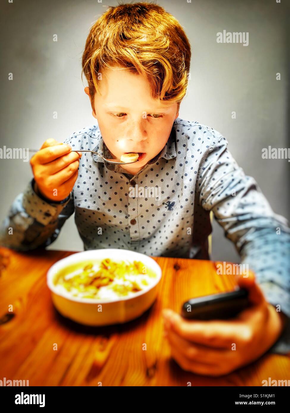 11-jährige junge Müsli zu essen, während der Blick auf Online-Inhalte auf einem Smartphone, Bawdsey, Suffolk, England. - Smartphone-aufgenommenes Stockfoto