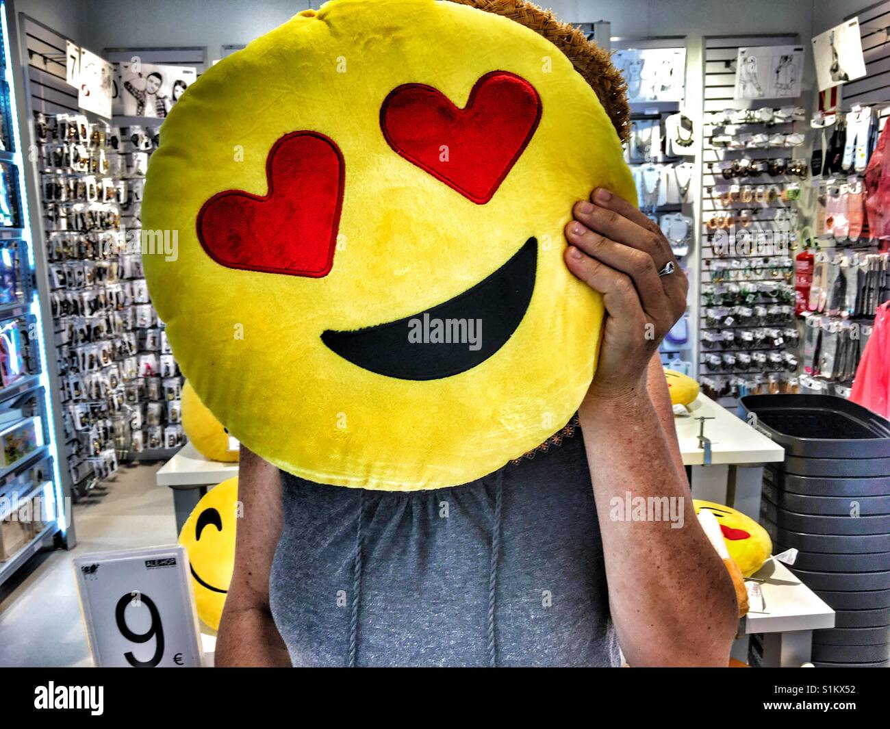 Frau mit einem I Love You Emoticon Kissen über ihr Gesicht in einem Geschäft - Smartphone-aufgenommenes Stockfoto