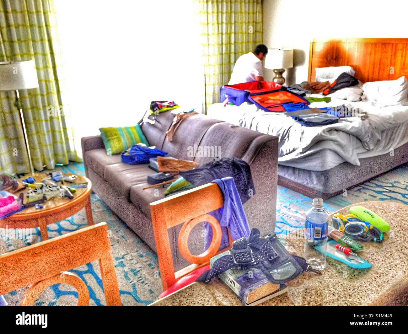 Packen Koffer im Hotel Zimmer am Ende der Ferien. Unordentlich Zimmer mit Elementen, die auf der Werkbank, Tisch und Lounge während Koffer auf das Bett und Person dahinter öffnen. Stockfoto