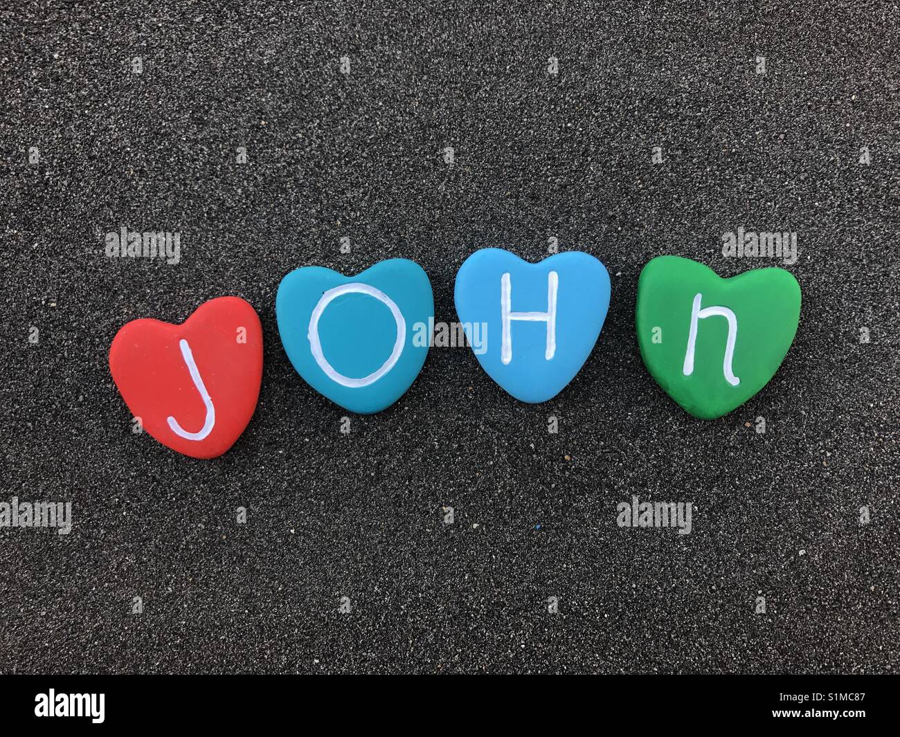 John, männlich Name mit bunten Herzen Steine über schwarzen vulkanischen Sand Stockfoto