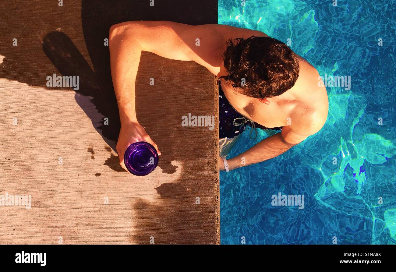 Junger Mann mit Getränk in der Hand am Rande einer Außenpool an einem heißen sonnigen Tag. - Smartphone-aufgenommenes Stockfoto
