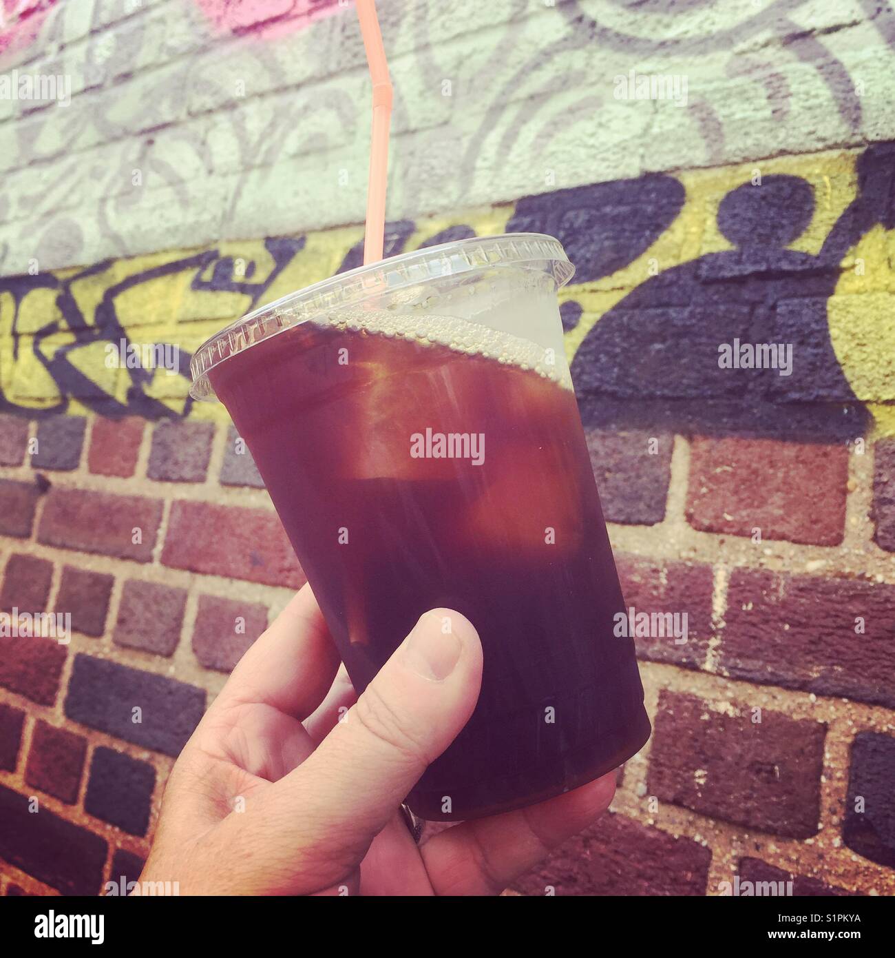 Eine Frau hält ein Glas Eiskaffee vor einer Ziegelmauer mit Graffiti drauf - Smartphone-aufgenommenes Stockfoto