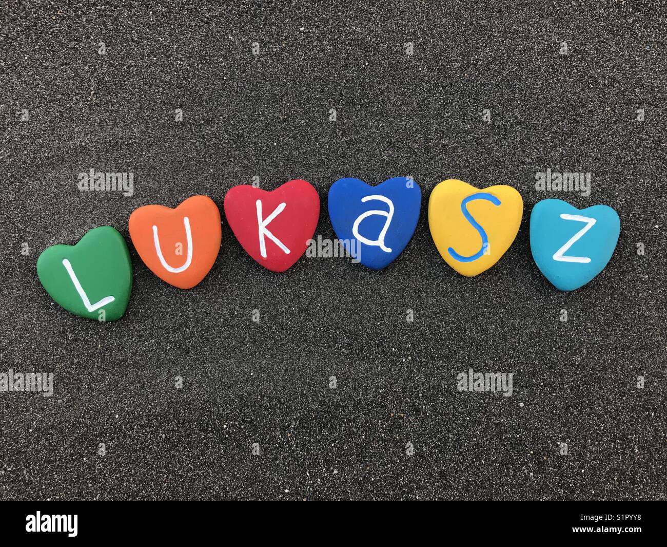 Lukasz, Polnisch männlicher Name mit bunten Herzen Steine über schwarzen vulkanischen Sand Stockfoto