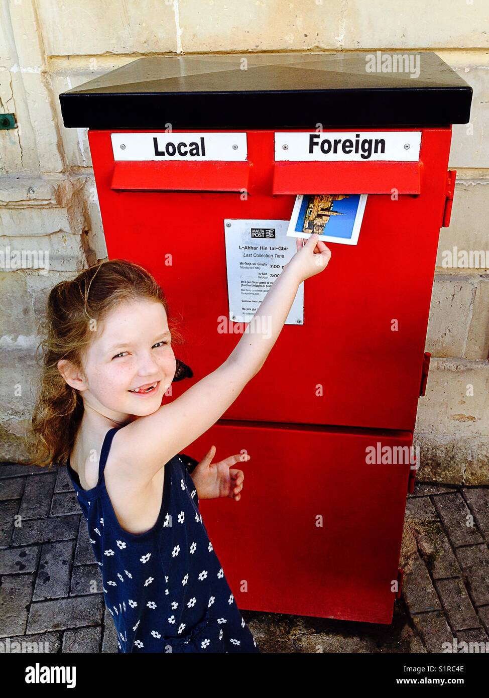 Kind/Kind/Mädchen eine Postkarte in den Briefkasten mit einem ausländischen Ziel gekennzeichneten Steckplatz ein. Malta. Stockfoto