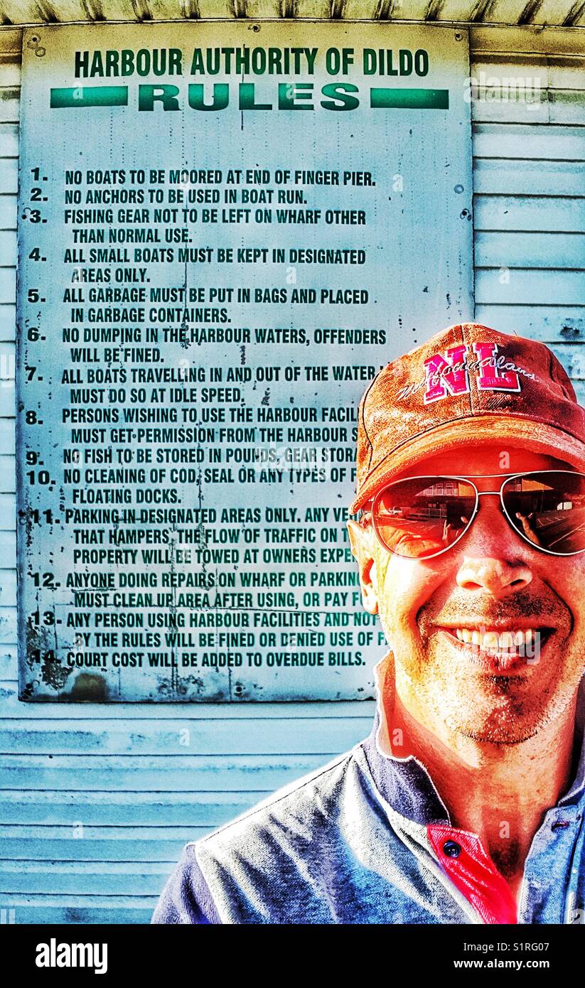 Lächelnder Mann mittleren Alters vor den Regeln der Hafenbehörde von Dildo, Dildo, Neufundland, Kanada. Dildo hat für seinen Namen Bekanntheit erlangt. - Smartphone-aufgenommenes Stockfoto