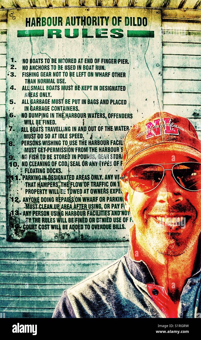 Lächelnder Mann mittleren Alters vor den Regeln der Hafenbehörde von Dildo, Dildo, Neufundland, Kanada. Dildo hat für seinen Namen Bekanntheit erlangt. - Smartphone-aufgenommenes Stockfoto