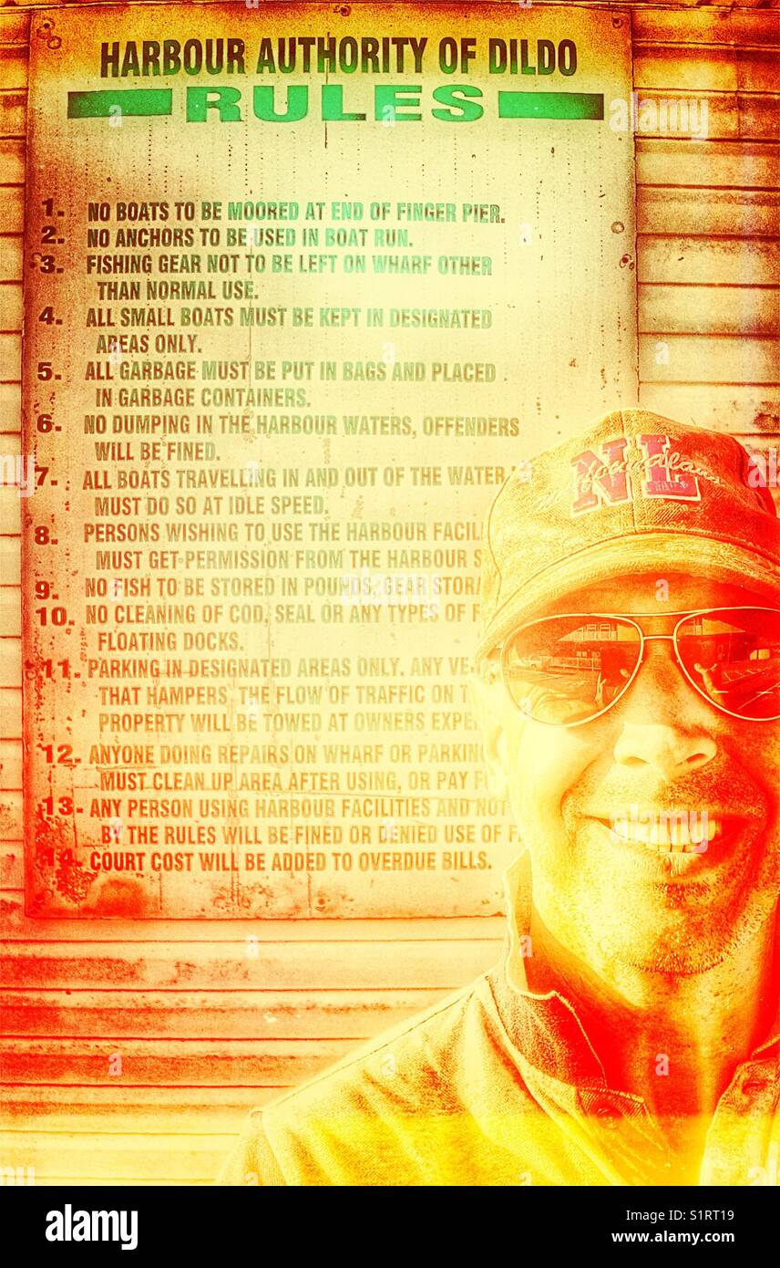 Lächelnder Mann mittleren Alters vor den Regeln der Hafenbehörde von Dildo, Dildo, Neufundland, Kanada. Dildo hat für seinen Namen Bekanntheit erlangt. - Smartphone-aufgenommenes Stockfoto