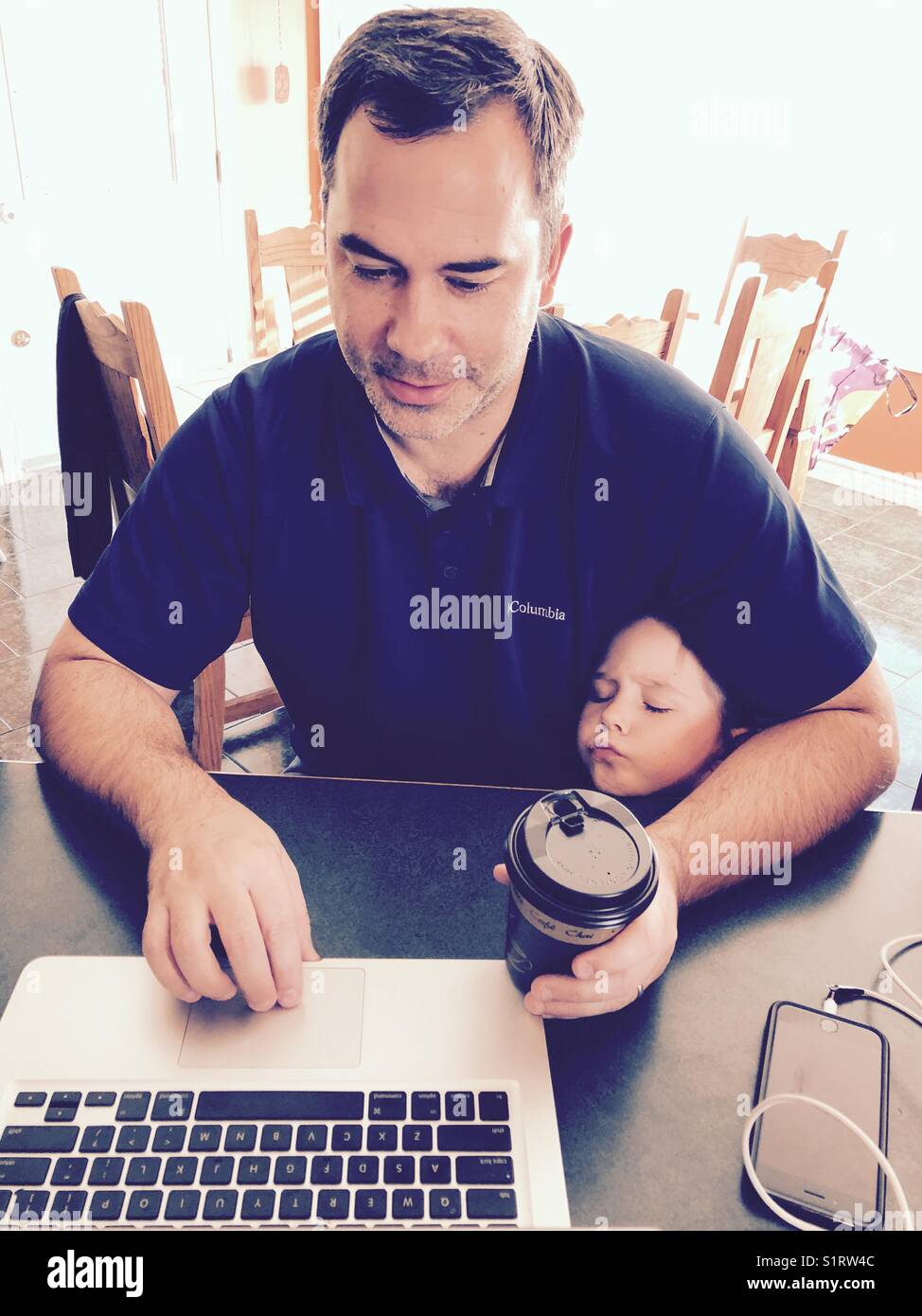 Mann mit Kaffee auf Laptop mit Tochter peeking unter seinem Arm für Aufmerksamkeit Stockfoto