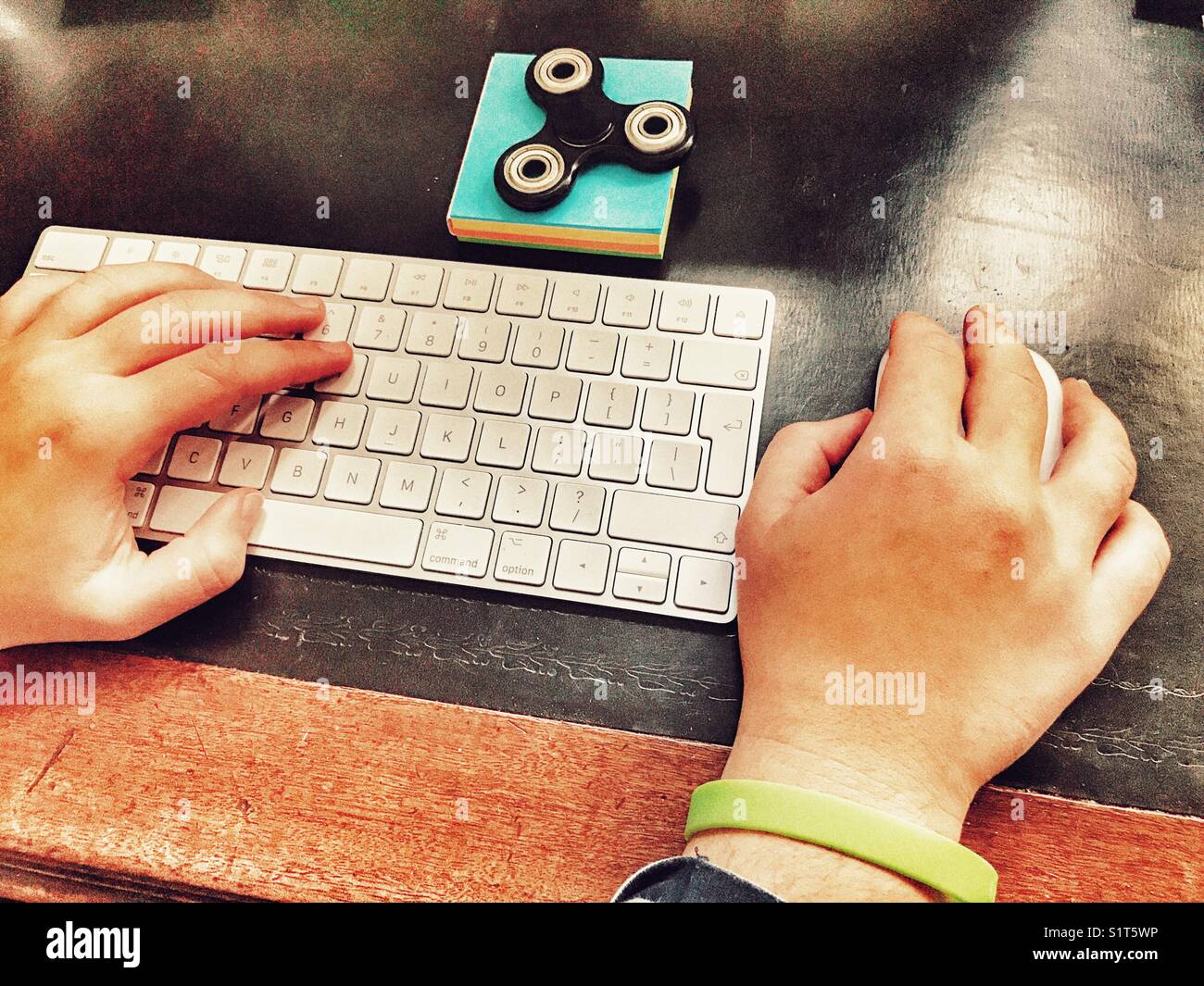 POV-persönliche Perspektive, Hände der Millennial-Frau auf Computertastatur und Maus mit einem zappelförmigen Spinner auf einem blauen Notizblock. - Smartphone-aufgenommenes Stockfoto