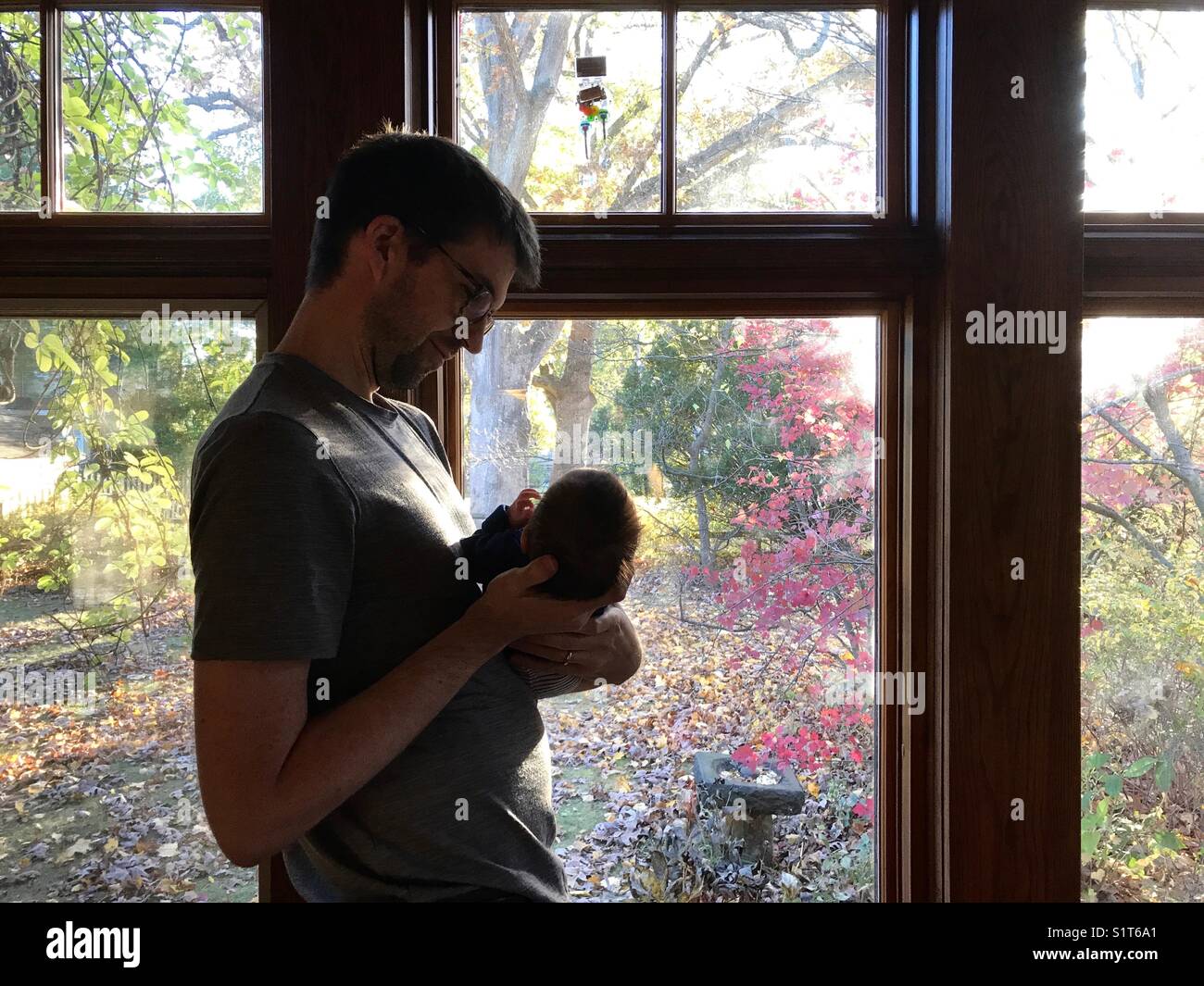 Ein Vater in den 30ern hält ein neugeborenes Baby, das neben einem Fenster in New Jersey, USA, steht. - Smartphone-aufgenommenes Stockfoto