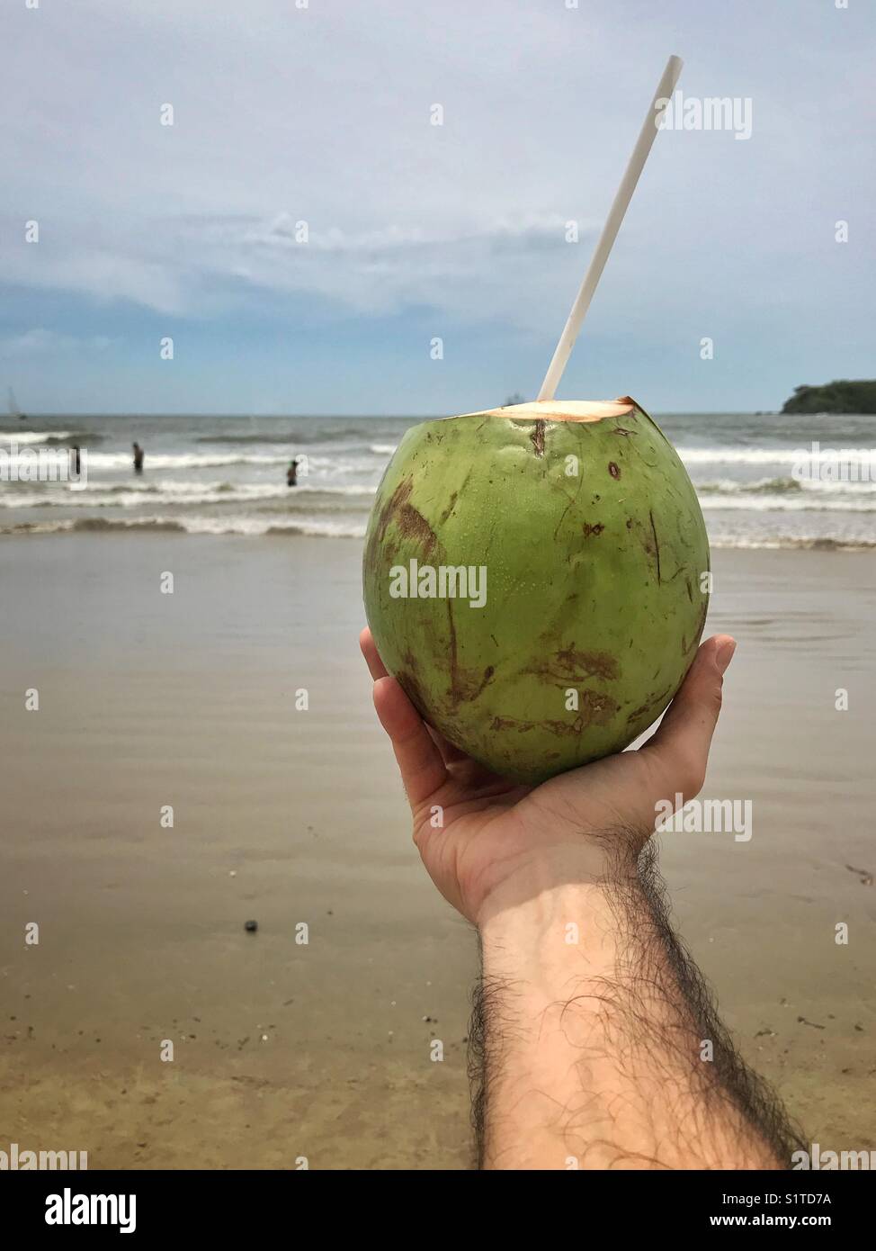 Die Hand des Mannes hält eine Kokosnuss am Strand vor dem Ozean im Hintergrund - Smartphone-aufgenommenes Stockfoto