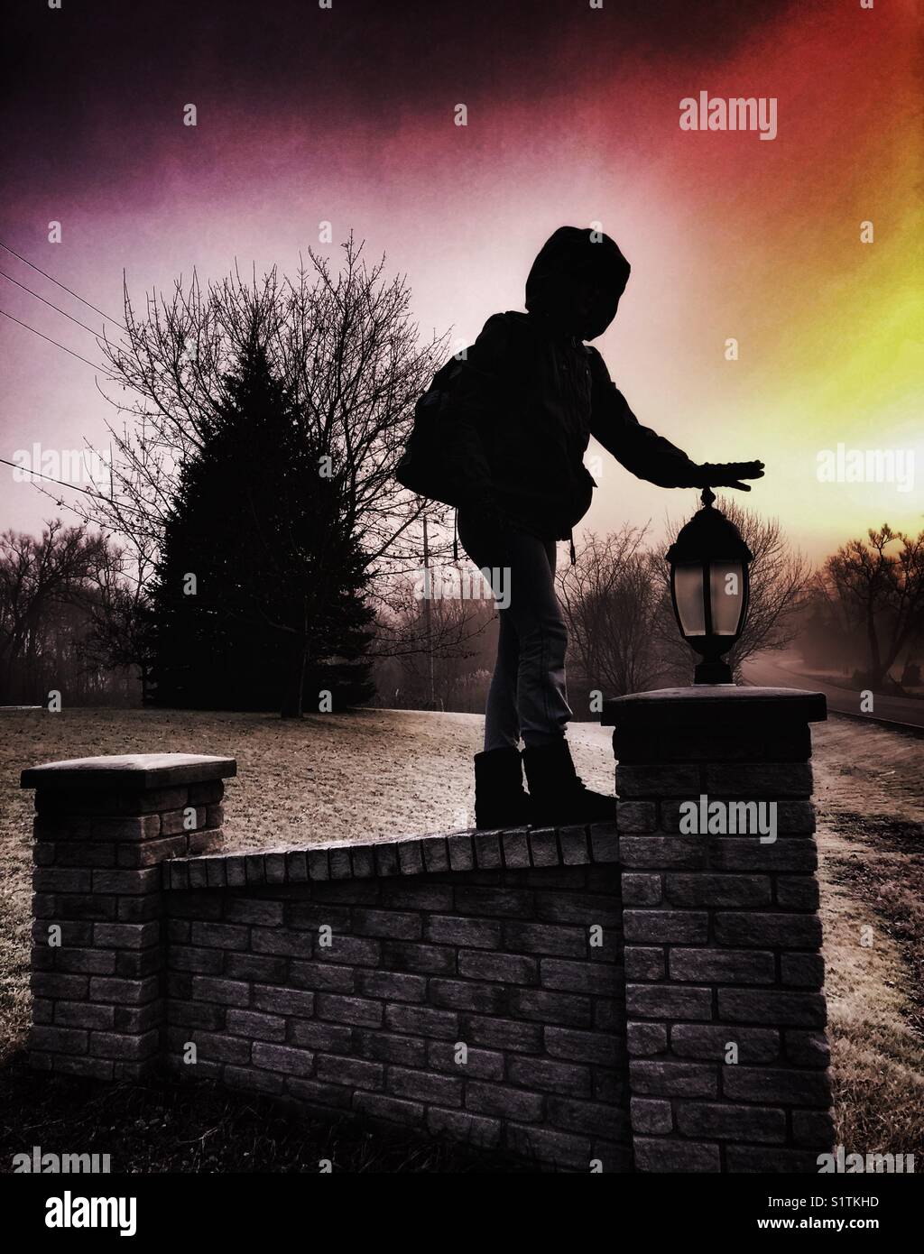 Die Silhouette eines Kindes, das an einer Ziegelwand balanciert und die Hand auf die Säulenlaterne legt - Smartphone-aufgenommenes Stockfoto