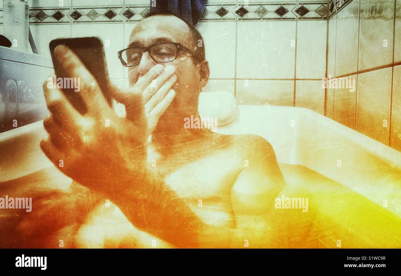 Fröhlich lachend im mittleren Alter Mann liegt in der Badewanne Blick auf Smartphone - Smartphone-aufgenommenes Stockfoto