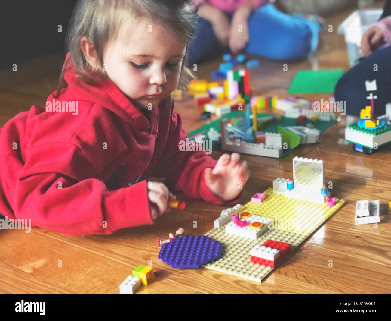 Kleinkind Mädchen in roten Kapuzenpulli Verlegung auf Holzboden LEGO spielen mit zwei anderen Kindern teilweise in Aussicht im Hintergrund - Smartphone-aufgenommenes Stockfoto