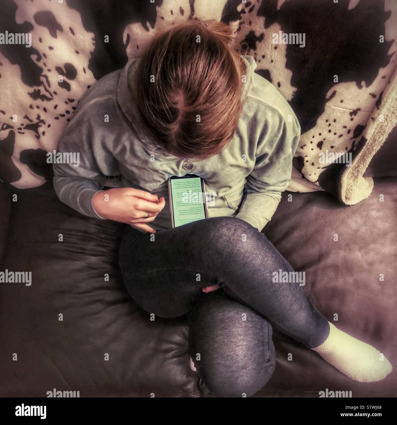 Von oben Blick auf Mädchen lesen auf dem Smartphone sitzen auf leder Sofa mit gekreuzten Beinen - Smartphone-aufgenommenes Stockfoto