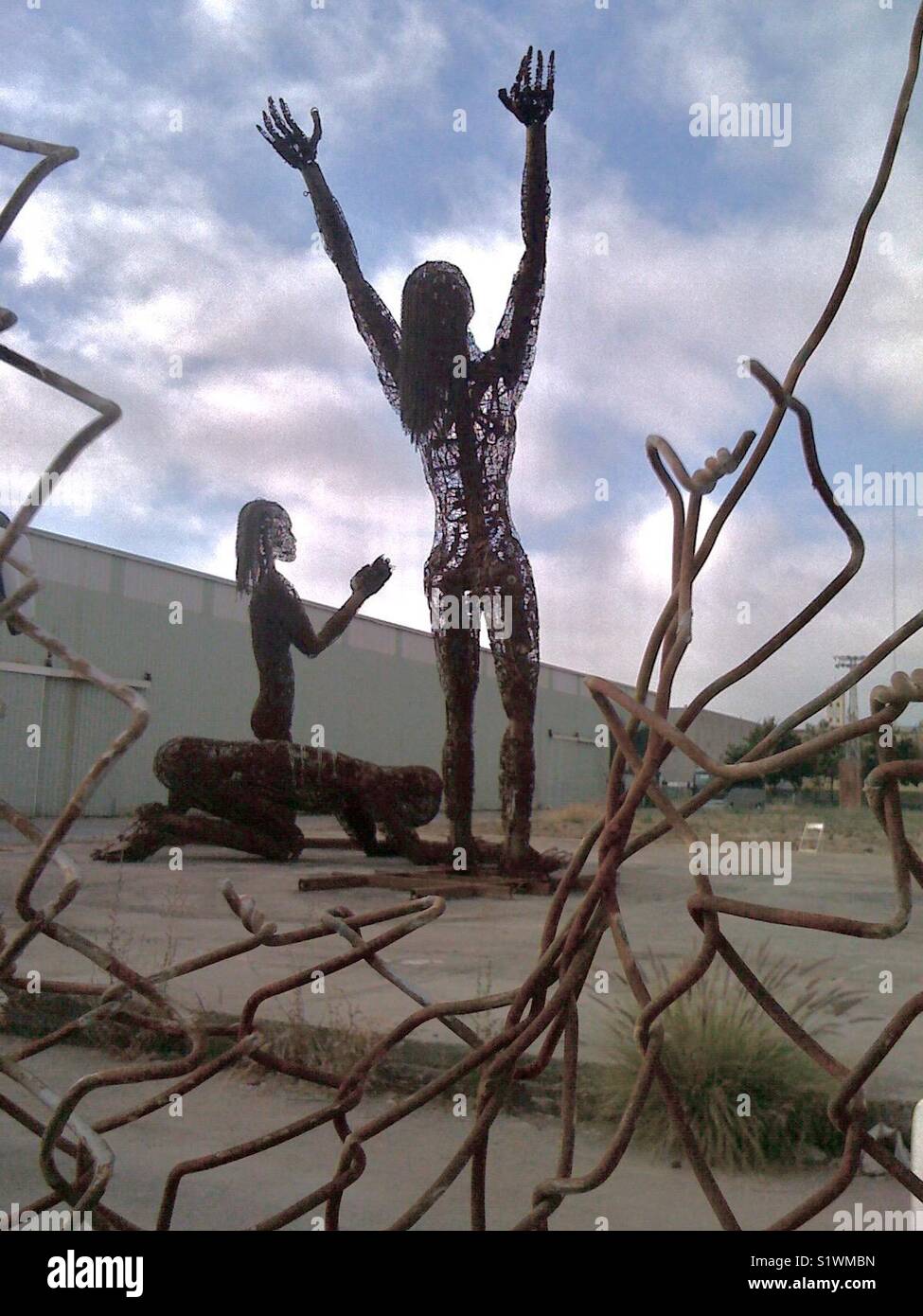 Stahlskulpturen von drei Betenden auf einem leeren Grundstück hinter gebrochenem Kettenglied, West Oakland, Kalifornien, USA. - Smartphone-aufgenommenes Stockfoto