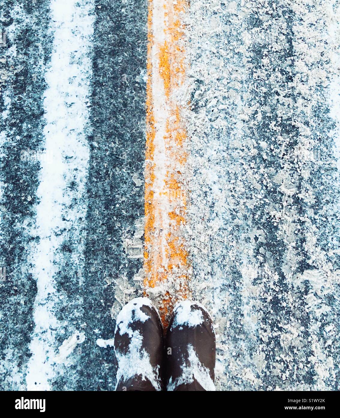 Blick nach unten an der braunen Stiefel der Person, die auf die gelbe Linie in der Mitte der verschneiten Piste - Smartphone-aufgenommenes Stockfoto