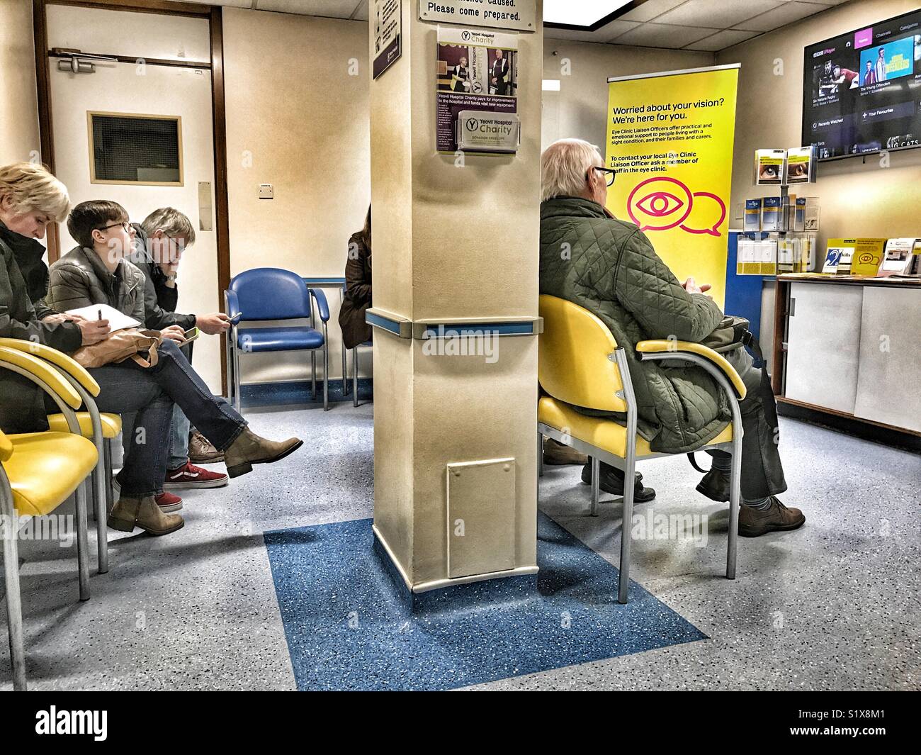 Die Menschen in den Wartebereich mit einem großen TV-Bildschirm, der Augenabteilung des NHS-Krankenhaus, Yeovil, Somerset, England Stockfoto