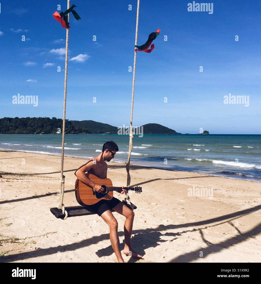 Ein Mann spielt Gitarre, während auf einer Schaukel sitzend genießen Sie den Strand und das Meer, während auf einer Insel, Koh Mak, Thailand Stockfoto