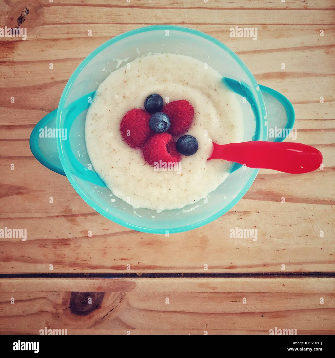 Kunststoff blau toddler Schüssel voller heißer Weizen Getreide, Himbeeren und Heidelbeeren mit einem roten Plastiklöffel auf einem Holztisch Stockfoto