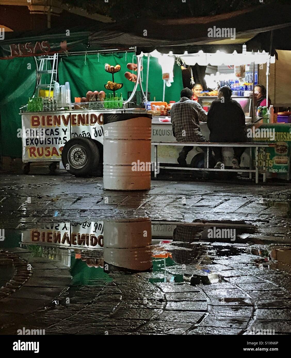 Ein Street Food vendor ist auch in einer Pfütze in einer regnerischen Nacht in Tlaquepaque, Mexiko, wo die Menschen sind, genießen Sie die köstlichen Speisen und Ambiente wider. Stockfoto