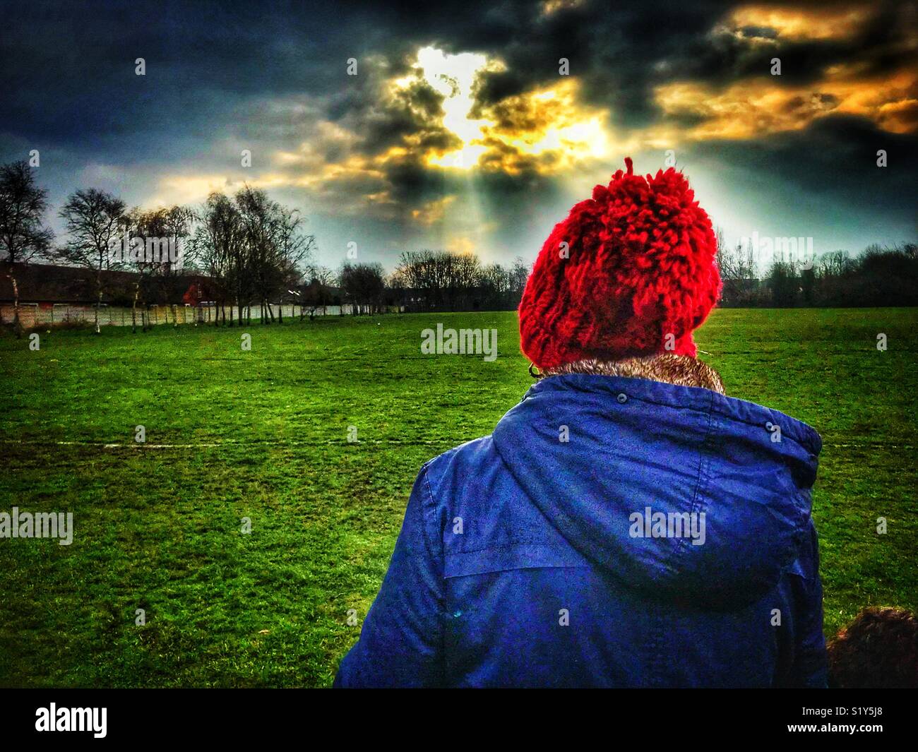 Dame mit einem blauen Mantel und Rot, aus Gewirken bobble hat zu Fuß in Richtung der frühen Morgensonne in einem Park in Warrington - Smartphone-aufgenommenes Stockfoto
