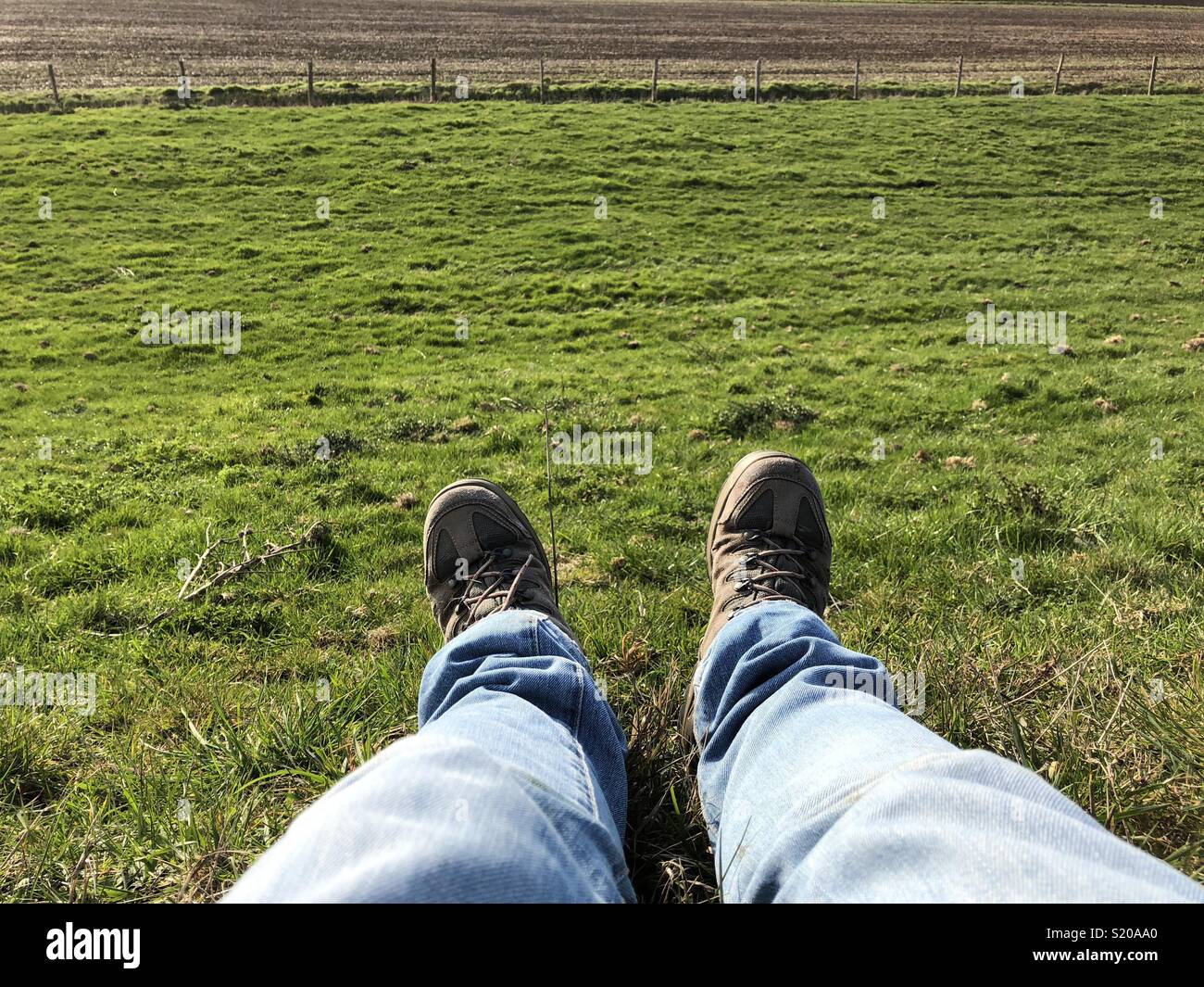 Mann Wanderer sitzen auf Gras mit Fokus auf die Beine trägt blaue Jeans und Wanderschuhe mit grünem Feld Wiese im Hintergrund Stockfoto