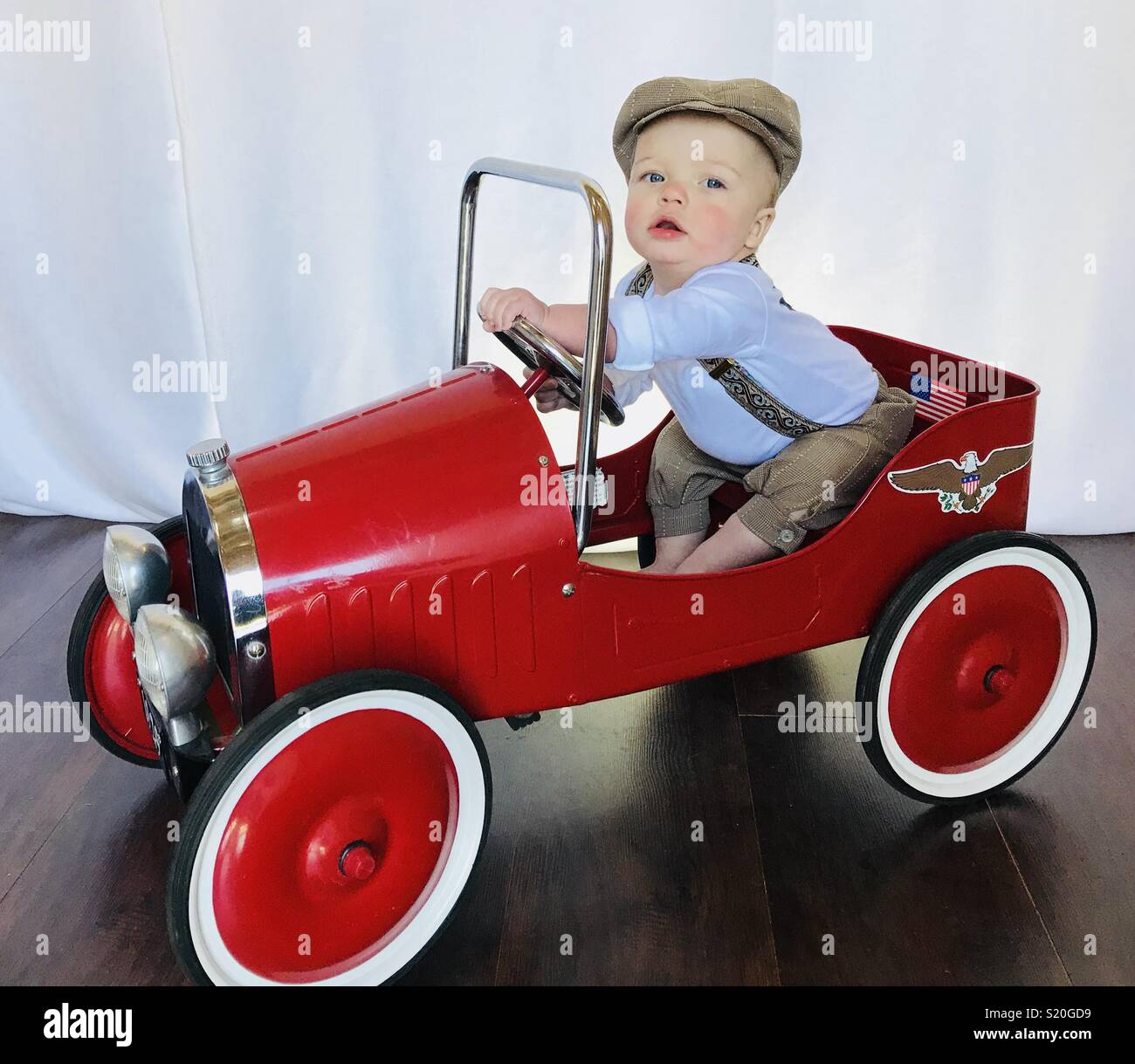 Cute Baby in einem Aktuelles boy Outfit spielen mit seinen antiken Auto  feilbieten. Diese niedlichen Baby trägt Braun tan Schlüpfer, Hosenträger  und ein aktuelles Boy hat, während er in seinem roten Auto