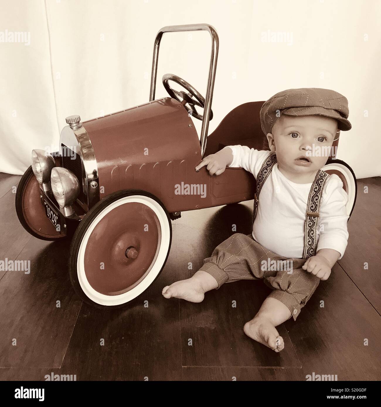 Cute Baby in einem Aktuelles boy Outfit spielen mit seinen antiken Auto feilbieten. Diese niedlichen Baby trägt Braun tan Schlüpfer, Hosenträger und ein aktuelles Boy hat während neben seinem Auto sitzen. Stockfoto