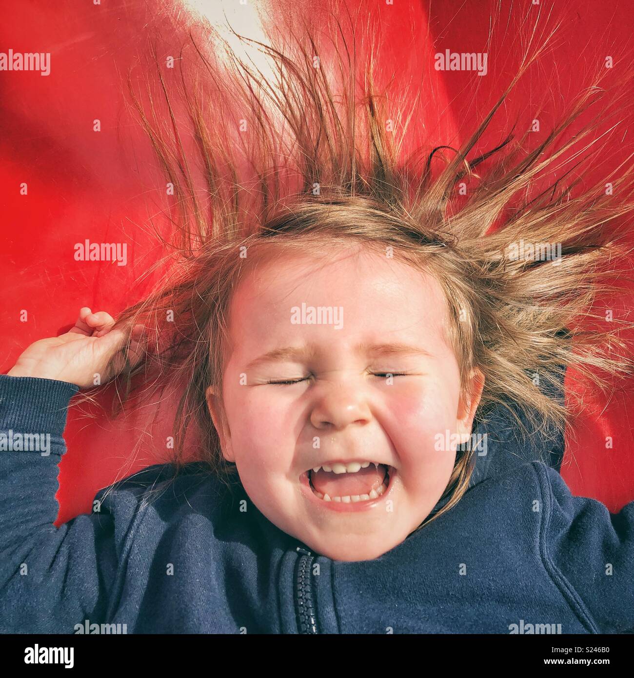 Portrait von Happy toddler girl Gesicht, während sie rutscht eine rote Folie mit geschlossenen Augen, den Mund offen und Haar kleben von statischer Elektrizität - Smartphone-aufgenommenes Stockfoto