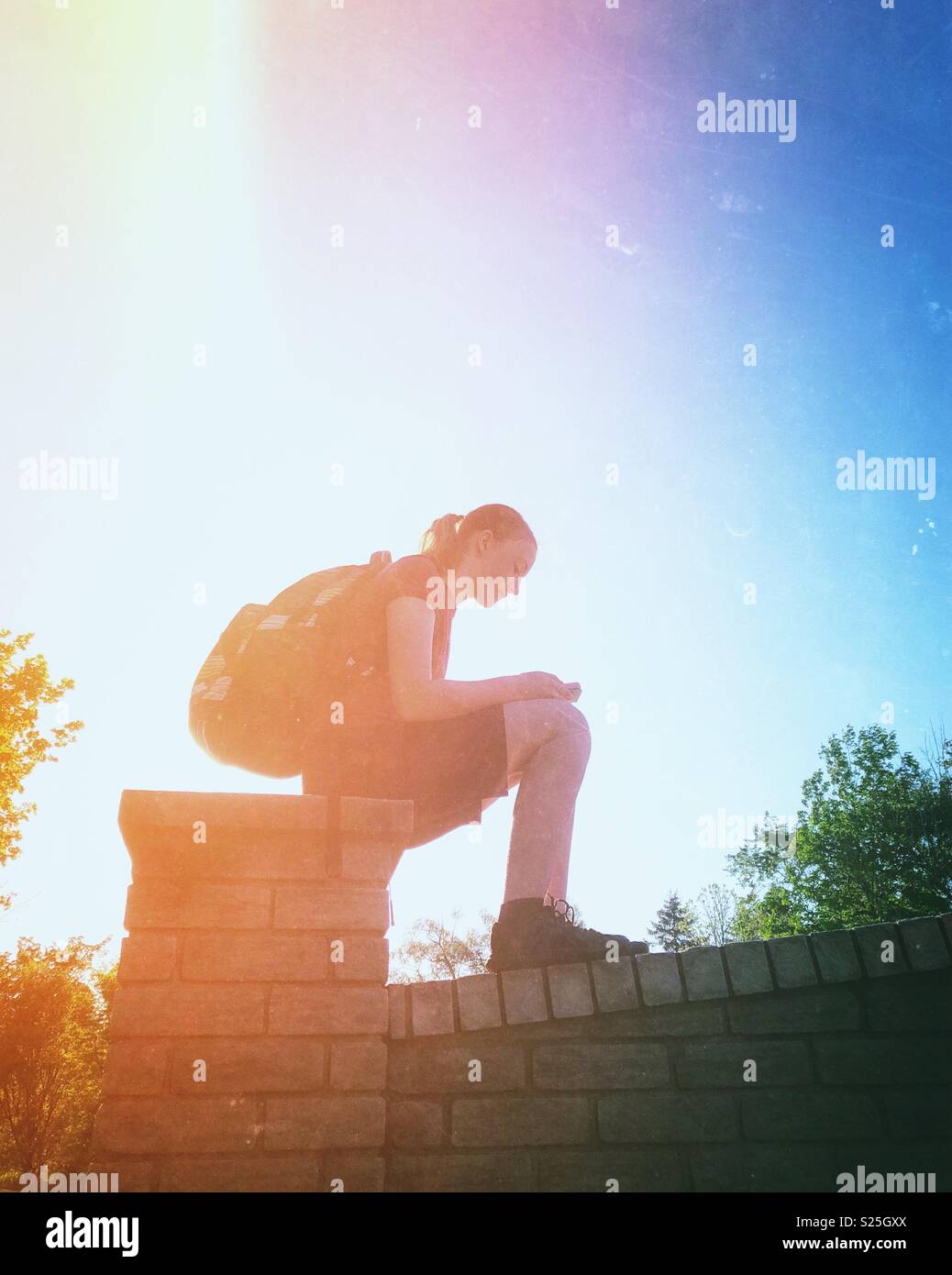 Auf der Suche nach einer Schülerin trägt einen Rucksack sitzt auf einem Stein Säule an Smartphone suchen mit morgen Licht im Hintergrund - Smartphone-aufgenommenes Stockfoto