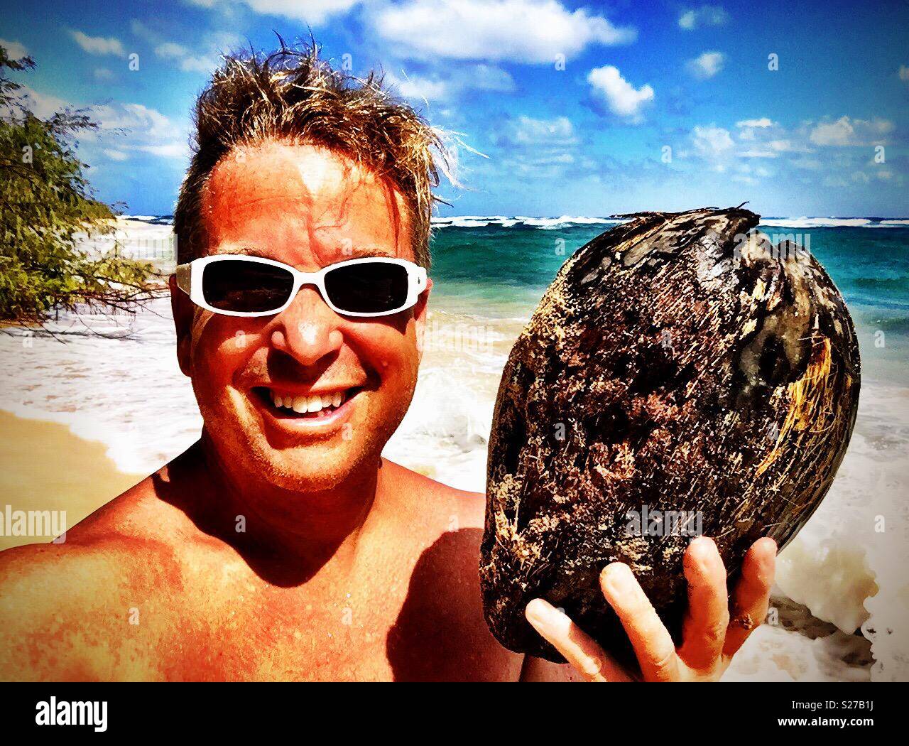 Vergnügtes gegerbt lächelnden Mann mit weissen Sonnenbrille sieht die Kokosnuss Geschenk vom Meer Stockfoto