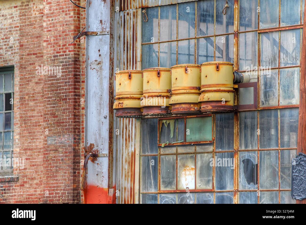 In diesem Foto einer verfallenen und verlassenen Fabrik, vier Rusty gelbe Fässer hang Hängen auf einer alten Fenster. Der Fensterrahmen ist rostig auf dieser Ziegelgebäude. Stockfoto
