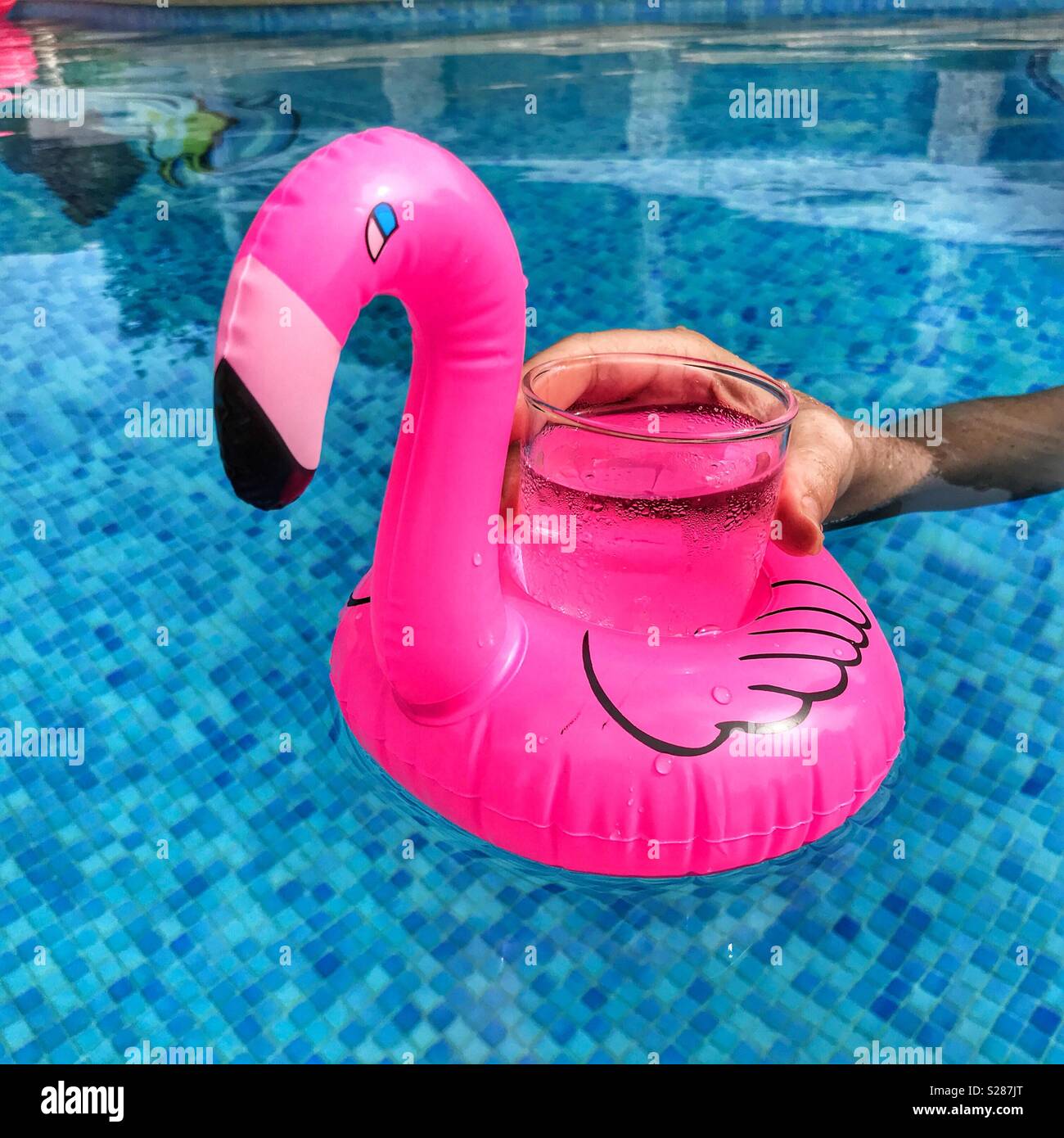 Woman's Hand erreichen für ein Glas in einem schwimmenden rosa Flamingo getränke Halter in einem Pool Stockfoto