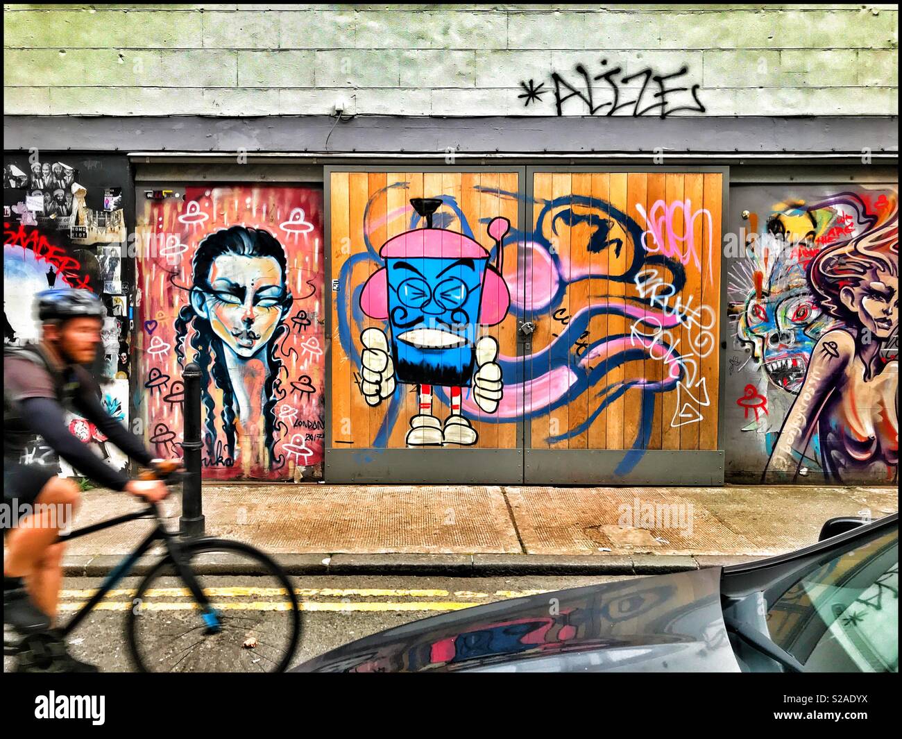 Ein Radfahrer fährt vorbei an einer Reihe von Türen geschmückt mit Street Art. Sind die Spray gemalten Bilder fantastische, kreative und inspirierende? Vielleicht sind Sie ein Chaos & konnte Sachschaden betrachtet werden? Foto © CH - Smartphone-aufgenommenes Stockfoto