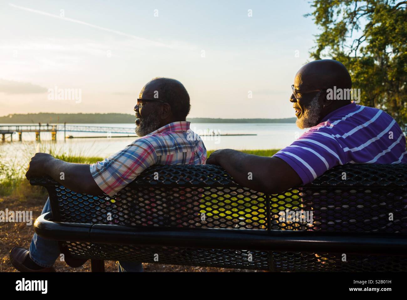 Zwei ältere Männer sitzen auf einer Bank an einem Sonnenuntergang über dem Wasser auf der Suche Stockfoto