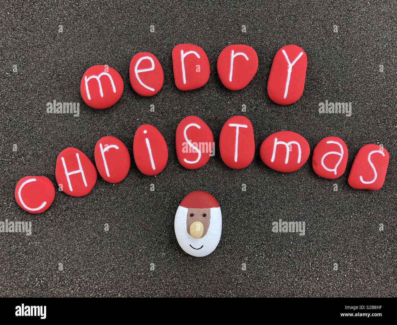Frohe Weihnachten Text mit rot gefärbten Steine, Santa Claus Fläche auf einem Stein über schwarzen vulkanischen Sand - Smartphone-aufgenommenes Stockfoto