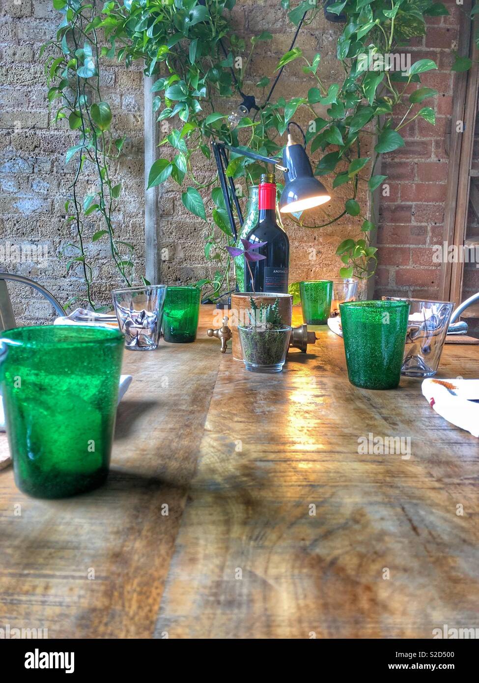 Tabelle der Einstellungen für das Mittagessen mit grünen tumbler Gläser, Rotwein und Zimmerpflanzen auf hölzernen Tisch gegen freiliegenden Ziegeln im Loft Stil industrielle Innenraum Stockfoto