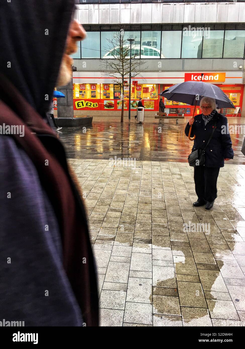 Eine ältere Frau mit Sonnenschirm, die alleine stehen im Regen, und ein Mann in die Szene Nahaufnahme von Links. Stockfoto