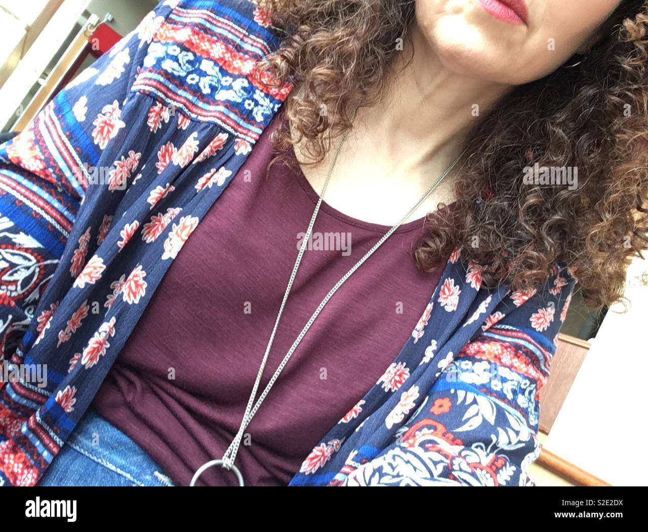 Curly dunkelhaarige Frau mit Silber Halskette gegen Burgund t-shirt rot und blau gemusterten fließende Jacke und tragen rosa Lippenstift Stockfoto