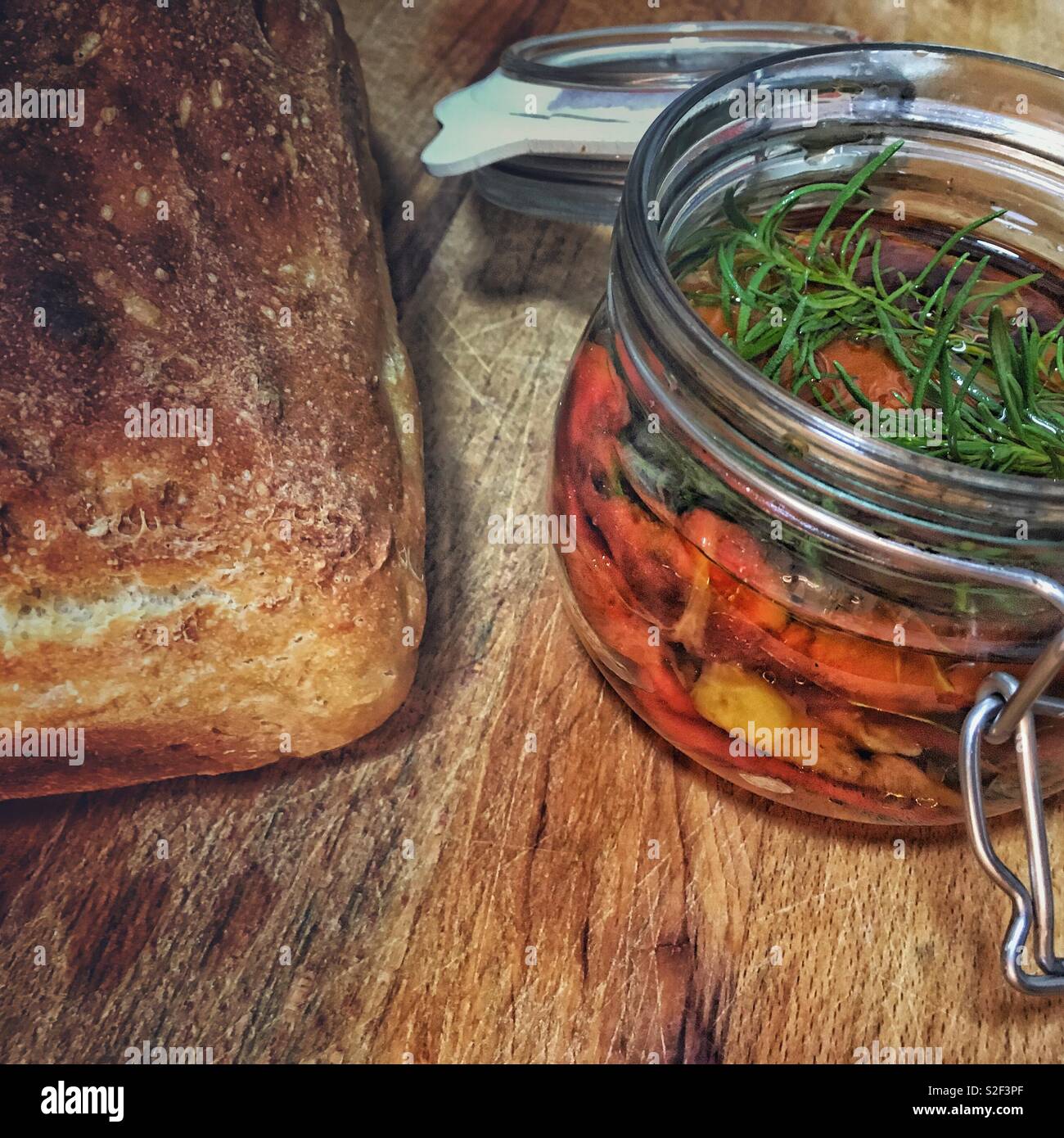 Ein hausgemachtes Brot und ein Glas hausgemachte marinierte Rote und gelbe Paprika mit einem Zweig Rosmarin an der Spitze. - Smartphone-aufgenommenes Stockfoto