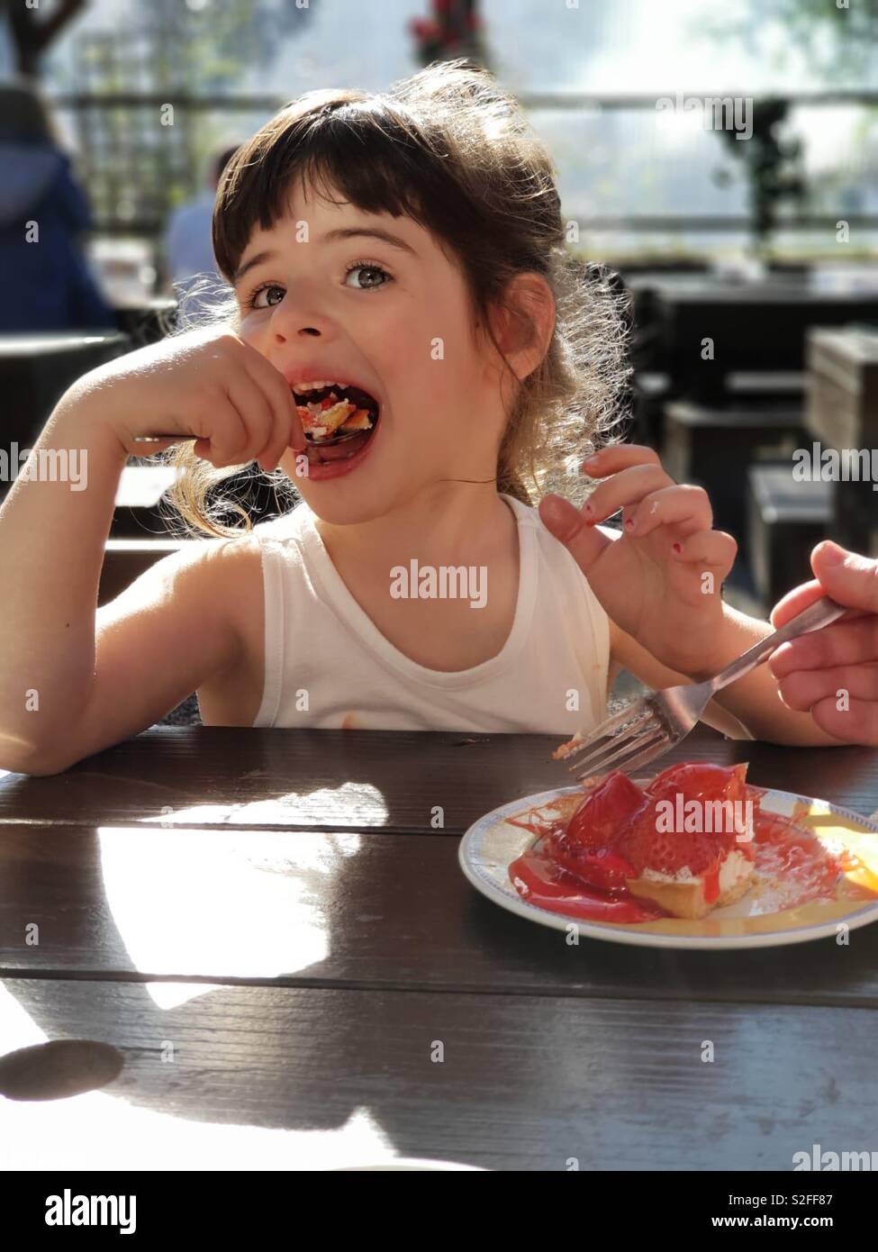 Vier Jahre alte nett schön Mädchen in einem weißen Weste essen einen Erdbeerkuchen mit großer Freude, gemeinsam mit Vater/Großvater. Große Augen, grosse Lippen, Süßes, Pudding, Datum, Wüste Stockfoto