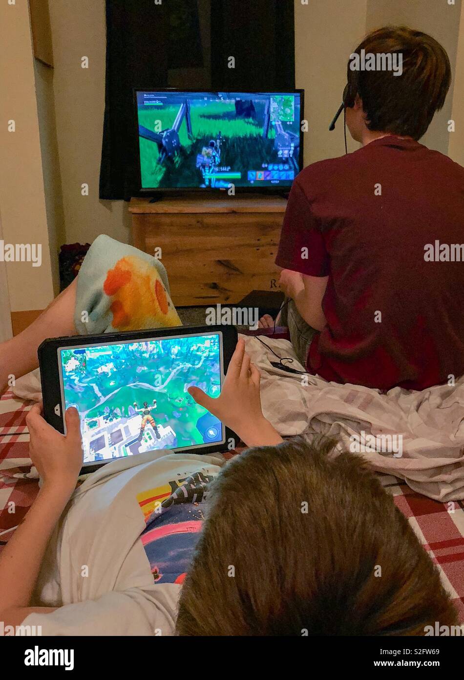 Zwei Brüder spielen Fortnite auf Ihrem eigenen Gerät. Der ältere Bruder  spielt auf den Fernseher im Hintergrund und der jüngere Bruder spielt auf  seinem Tablet im Vordergrund Stockfotografie - Alamy