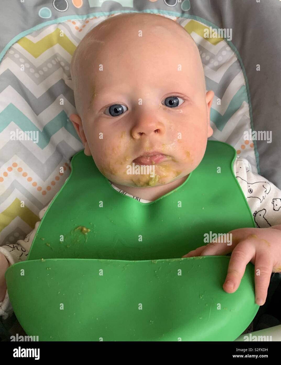 Baby Junge mit blondem Haar und blauen Augen im Hochstuhl sitzen mit grünen Erbsen alle über Gesicht. Eine grüne Bib. - Smartphone-aufgenommenes Stockfoto
