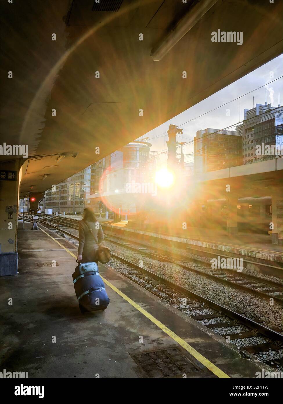 Eine nicht erkennbare junge Frau, die ihr Gepäck über die Plattform von einem Bahnhof in einer hellen und farbenfrohen Halo von Licht der untergehenden Sonne abgedeckt Stockfoto