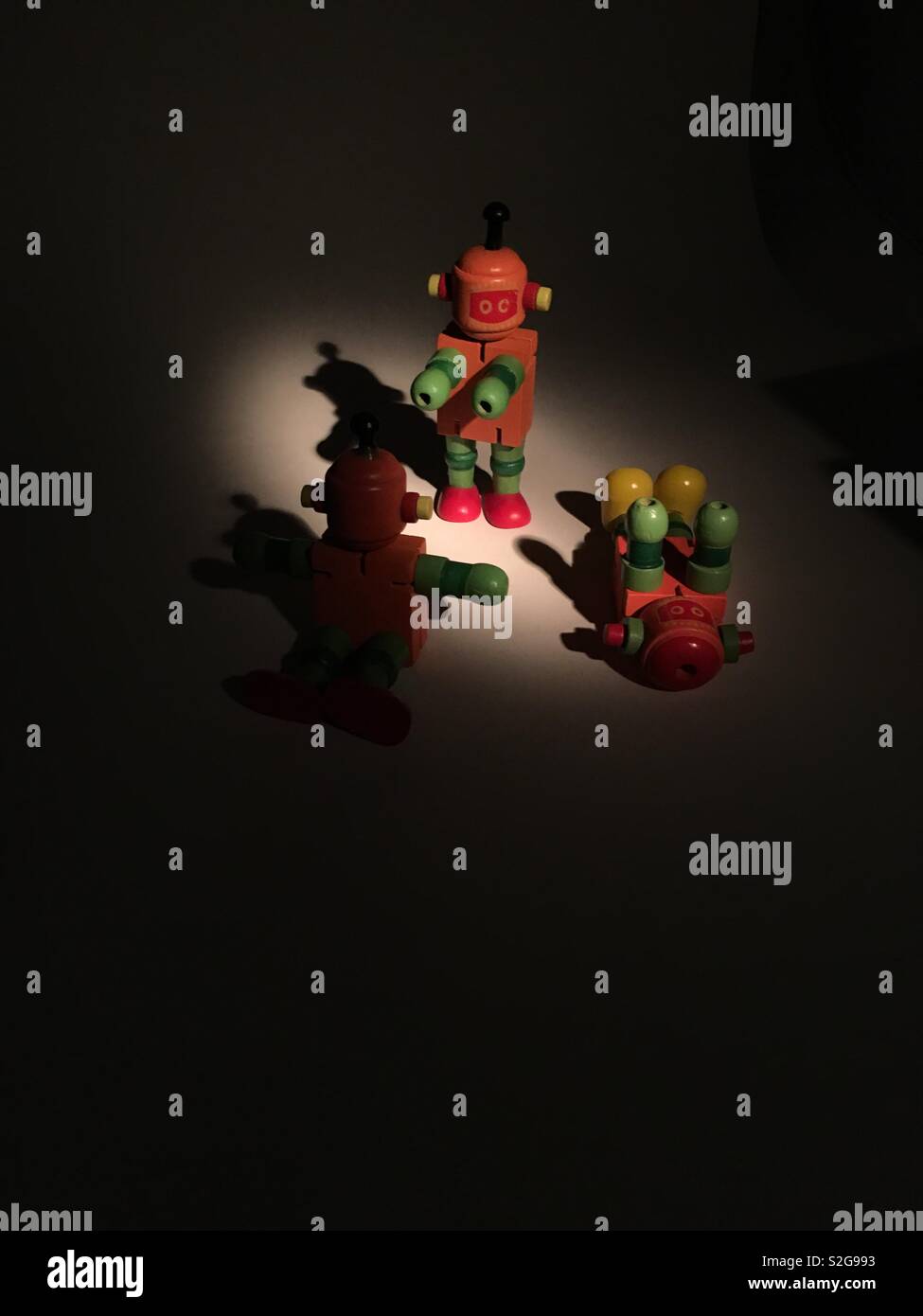 Drei Holz- orange und grün Spielzeug Roboter in einem Spotlight mit dunklen umgibt. Das eine ist stehend, kniend, und eines auf dem Rücken. Stockfoto